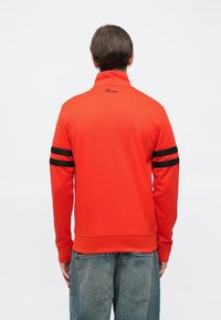 Punane lukuga jope mustade triipudega varrukaaladel, valmistatud tekstuurse materjaliga, millel on kõrge krae ja seljal tikandiga logo.