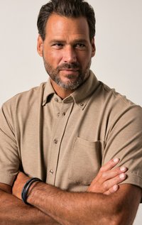 Beige overhemd met korte mouwen, een button-down kraag, borstzak en stofstructuur. Model heeft de armen gekruist en draagt een zwarte armband.