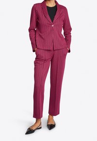 Fluorescerend roze en zwart patroonpak met een nauwsluitend colbert met een ingesneden kraag en een enkele knoopsluiting, gecombineerd met op maat gemaakte broek en platte schoenen.