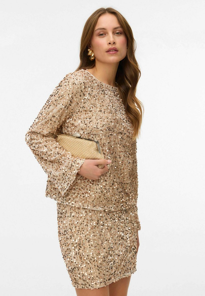 Vestido de cóctel con cuentas en tono nude, con mangas largas, que presenta un corte holgado y un diseño con capas, acompañado de un bolso beige y acentos dorados.