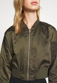 Olivově zelená bomber bunda s krátkým střihem, elastickými manžetami a zlatým zipem. Hladká, saténová textura.