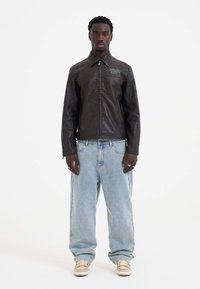 Brown leather jacket with a zip front and pointed collar, paired with light blue oversized jeans and light-colored sneakers.