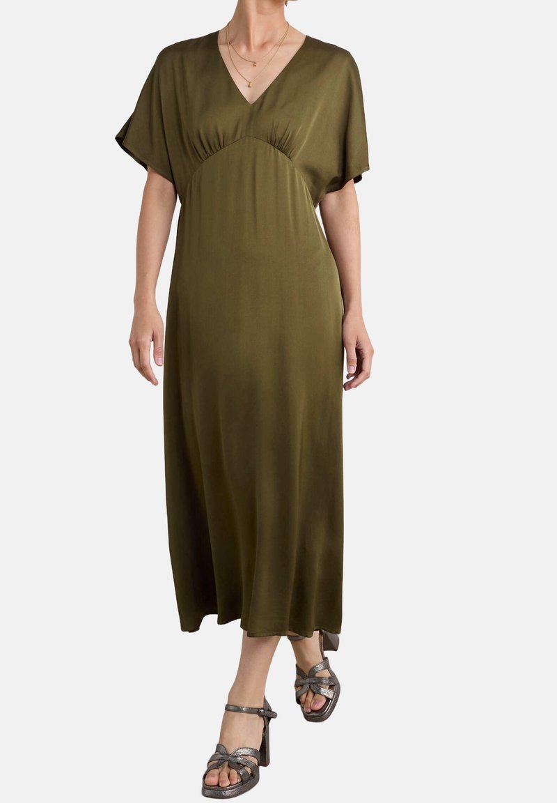Femme portant une robe midi vert olive à décolleté en V avec des manches courtes et des sandales métalliques à talons hauts, debout devant un fond uni.