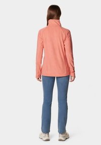 Femme debout, vue de dos, portant une veste polaire corail, un jean bleu et des baskets beiges sur fond blanc.