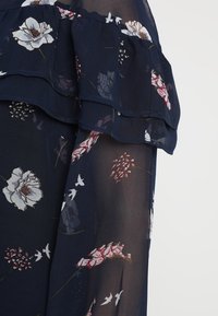 Dunkelblaue Bluse mit floralem Druck, ausgestattet mit geschichteten Rüschendetails. Hergestellt aus leichtem, transparentem Stoff mit Texturen, die Tiefe und Bewegung verleihen.