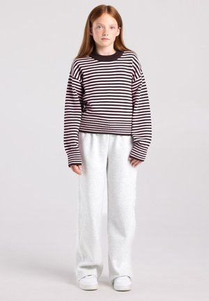 Fille aux longs cheveux roux portant un pull rayé noir et rose, un pantalon gris clair et des chaussures blanches, debout devant un fond uni.