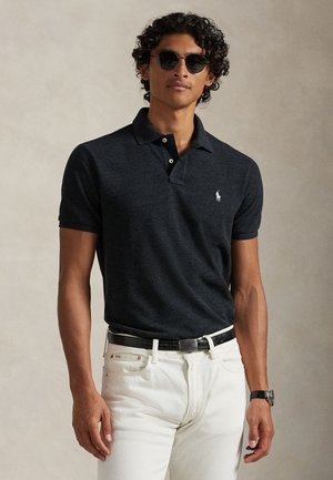 Jeune homme aux cheveux bouclés portant des lunettes de soleil noires, un polo foncé, un pantalon blanc, une ceinture noire et une montre-bracelet, debout devant un fond uni.