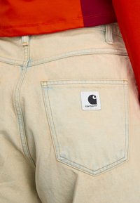 Ljusblå denimjeans med gula sömmar, med en rektangulär bakficka och en vit Carhartt-logotyp.