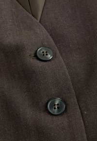 Braunstoff mit grober Textur, der zwei dunkle Knöpfe mit jeweils vier Löchern zeigt, die auf der Vorderseite einer Jacke angebracht sind.