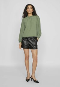 Groene blouse met hoge hals en lange mouwen, gecombineerd met zwarte leren shorts en puntige zwarte slingback hakken.