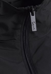 Juodas užtrauktukas striukė su tvirtu juodu užtrauktuku ir prekės ženklo traukimo lazdele su minimalistiniu logotipu. Audinys turi glotnų, matinį paviršių.