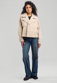 Beige jakku yang dipotong dengan lapel lebar dan tombol double-breasted, dipadukan dengan celana jeans longgar berwarna biru dan sepatu chunky hitam.