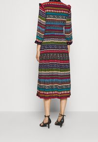 Robe longue multicolore avec des motifs horizontaux variés, dotée d'épaules froncées et de manches trois-quarts. Portée avec des talons noirs.