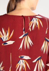 Bordeaux bluse med et blomsterdesign i krem, orange og blå. Bagsiden har en lille knaplukning og en nøglehulåbning. Glat tekstur.