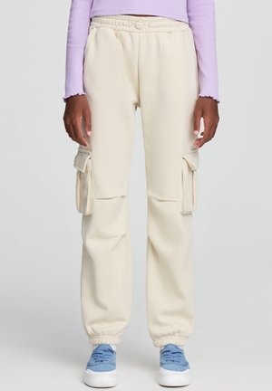 Pantaloni cargo beige realizzati in morbido materiale, dotati di tasche laterali, una vita elasticizzata e orli a costine, abbinati a sneaker azzurro chiaro.
