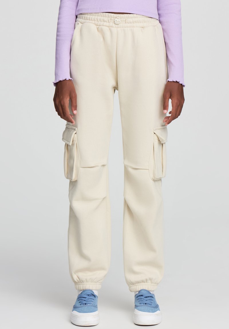 Beżowe spodnie cargo wykonane z miękkiego materiału, z bocznymi kieszeniami, elastycznym paskiem w talii i mankietami, zestawione z jasnoniebieskimi sneakersami.