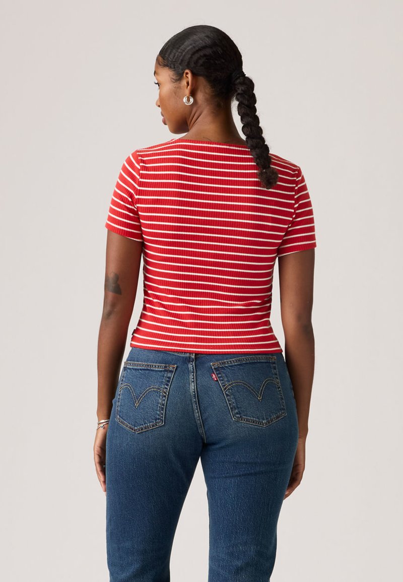 Femme aux cheveux tressés portant des boucles d'oreilles en argent en forme d'anneau, un haut à manches courtes rayé rouge et blanc, et un jean bleu en denim, vue de dos.