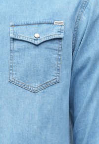 Gaiši zils džinsa krekls ar smailu krūšu kabatu, sudraba aizdares pogu un kārtīgu šuvumu. Audums šķiet gluds. Redzama zīmola etiķete.