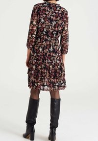 Robe à imprimé floral en tissu noir transparent avec manches longues, volants à étages et taille cintrée, associée à des bottes en cuir noir montantes jusqu'aux genoux.