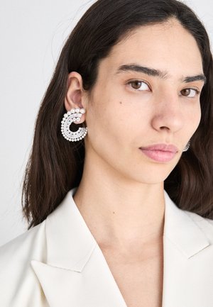 Große kreisförmige Ohrringe mit klaren Kristallen und Perlenakzenten, in einem schimmernden Silber-Finish, getragen von einem Model mit glattem Haar.