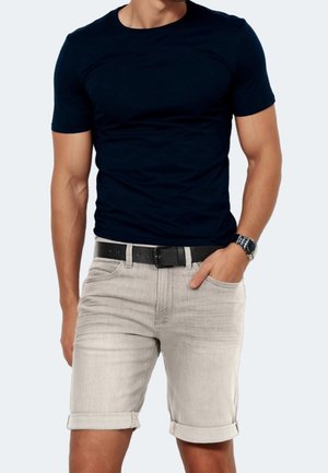 Homme portant un t-shirt ajusté bleu marine à manches courtes, un short en jean gris clair retroussé avec une ceinture noire, et une montre-bracelet au bras gauche.