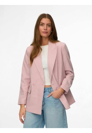 Femme aux longs cheveux bruns portant un blazer rose clair sur un haut blanc et un jean bleu clair, debout avec une main dans la poche.