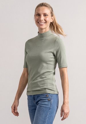 Femme souriante aux cheveux blonds attachés en queue de cheval, portant un pull à col roulé à manches courtes vert clair et un jean bleu, debout devant un fond uni.