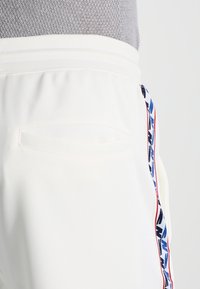 Vita svettbyxor med en texturerad midja, sidoficka och ett färgglatt mönstrat rand längs den yttre sömmen i blått, rött och svart.