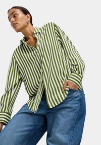 Camicia a maniche lunghe a righe verde e bianche con colletto button-down, abbinata a jeans di denim blu. Caratterizzata da un fit rilassato e polsini larghi.