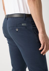 Chinos azul marinho com uma textura suave, apresentando um bolso traseiro com botão e um rótulo de logotipo. Um cinto de couro preto é visível na cintura.