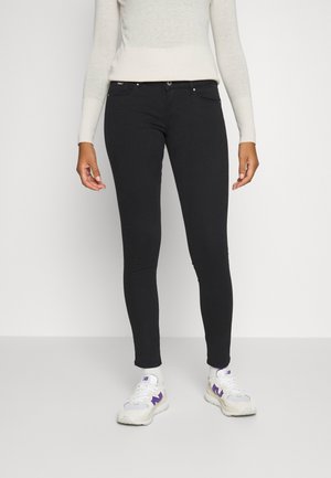 Jean skinny noir avec une texture lisse, présentant des poches avant, une fermeture à bouton et une coupe ajustée. Porté avec des baskets blanches.