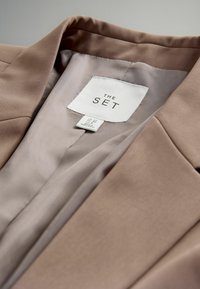 Blazer beige con forro de satén. Presenta un cuello con muescas, textura suave y etiqueta de la marca "THE SET" cosida en el interior.