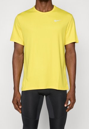 Mann trägt ein knallgelbes Nike-Sportshirt mit kurzen Ärmeln und schwarze enganliegende Sportleggings vor einem einfarbigen Hintergrund.