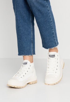 Persoon die witte hoge Palladium sneakers met beige zolen en blauwe cropped jeans draagt en op een grijze vloer staat.