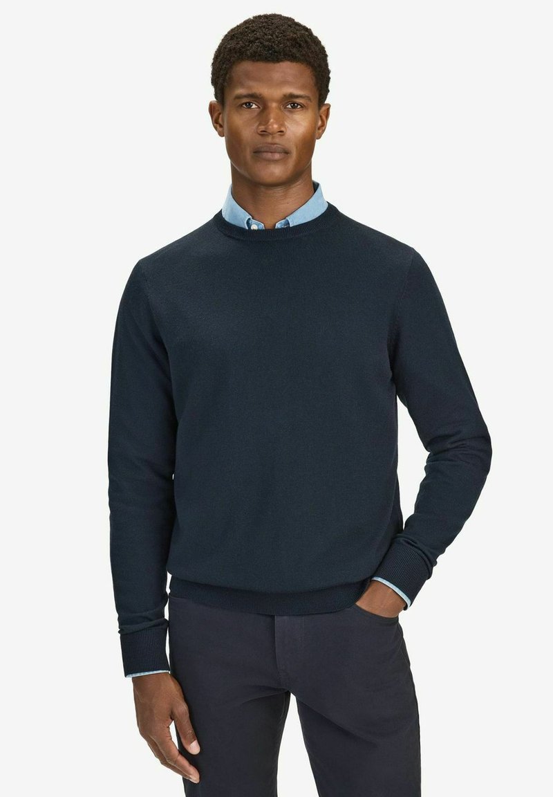 Mörkblå stickad tröja med rund halsringning och långa ärmar, med ribbade muddar och nederkant. Bärs över en ljusblå skjorta med krage.