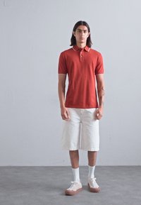 Sarkans polo krekls ar baltu apkakli, saskaņots ar baltām šortiem. Modelis valkā gaišas krāsas sporta apavus un baltas zeķes uz neitrāla fona.