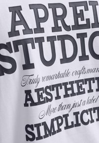 Valkoinen puuvillapaita, jossa on rohkeaa mustaa tekstiä. Avainlauseita ovat "APPRECIATE", "STUDIO" ja "SIMPLICITY", ja niissä on vaihteleva typografia.