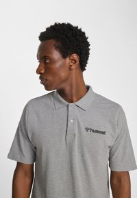Szara koszulka typu polo wykonana z miękkiego materiału, z dwoma guzikami u góry i czarnym logo "hummel" po lewej stronie na piersi. Krótkie rękawy.