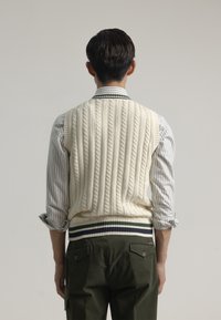 Chaleco de punto trenzado crema con cintura de canalé, llevado sobre una camisa a rayas con mangas remangadas. Unos pantalones cargo verde oscuro completan el look.