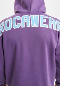 Lilla hettegenser med stor, lys blå "ROCAWEAR" tekst på baksiden. Materialet virker mykt med en glatt tekstur. Har hette.