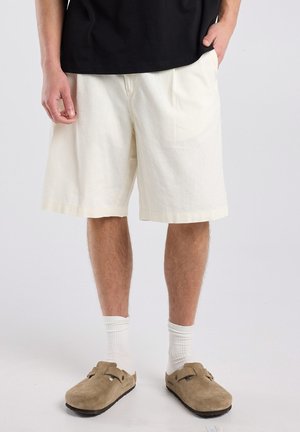 Man draagt beige korte broek, witte sokken en tan slip-on schoenen, staat met één hand in de zak tegen een effen achtergrond.