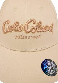 Casquette de baseball beige avec broderie "Calo Colucci" et "Milano 1973" en rose clair. Présente une étiquette ronde brillante avec des accents noirs et pastel.