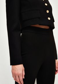 Zwarte cropped blazer met gouden knopen, vervaardigd uit een textuurstof en voorzien van klepzakken, gecombineerd met hoge zwarte leggings.