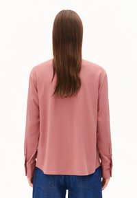 Person with long brown hair wearing a loose pink long-sleeve blouse and blue jeans, facing away against a plain white background.