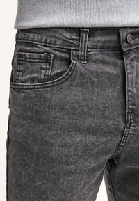 Jeans de denim gris oscuro que cuentan con un diseño de cinco bolsillos, con una textura ligeramente deslavada y costuras distintivas a lo largo de las costuras y bolsillos.