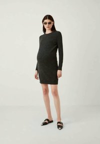 Fekete, hosszú ujjú ruha, testhez simuló sziluettel, texturált anyagból készült, térdig érő, fekete slip-on cipővel viselve, fémes díszítésekkel.