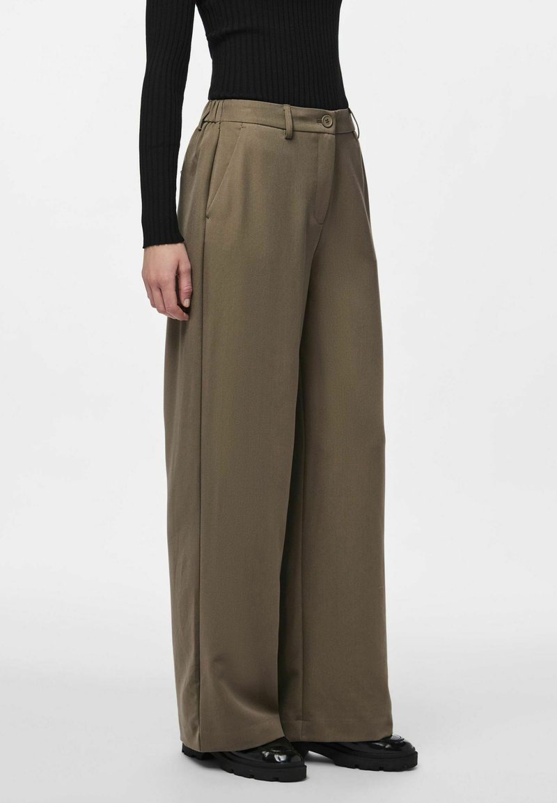 Femme portant un pantalon large olive et un haut noir côtelé à manches longues, debout avec les mains détendues le long du corps, chaussures noires épaisses visibles.