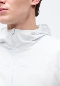 Κοντινό πλάνο ενός ατόμου που φοράει μια ανοιχτό γκρι ζακέτα The North Face με κουκούλα, εμφανίζοντας το στήθος, τον λαιμό και το κάτω μέρος του προσώπου.