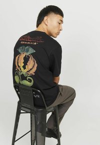 Černé tričko s barevným designem dýně a grafickým očima na zadní straně. Model sedící na černém kovovém stoličce, v šedých kalhotách.