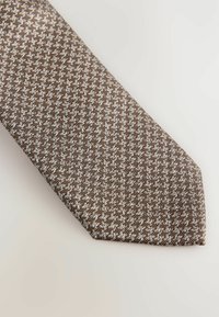 Ruskea solmio, jossa on houndstooth-kuvio vaihtelevilla vaaleilla ja tummilla sävyillä. Se on valmistettu teksturoidusta kankaasta, ja siinä on terävä pää.